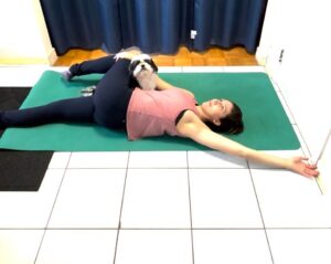 Sasha demonstrating the twisted angel by lying on her back, hips twisted in a 4 position, slowly stretching 1 arm out overhead